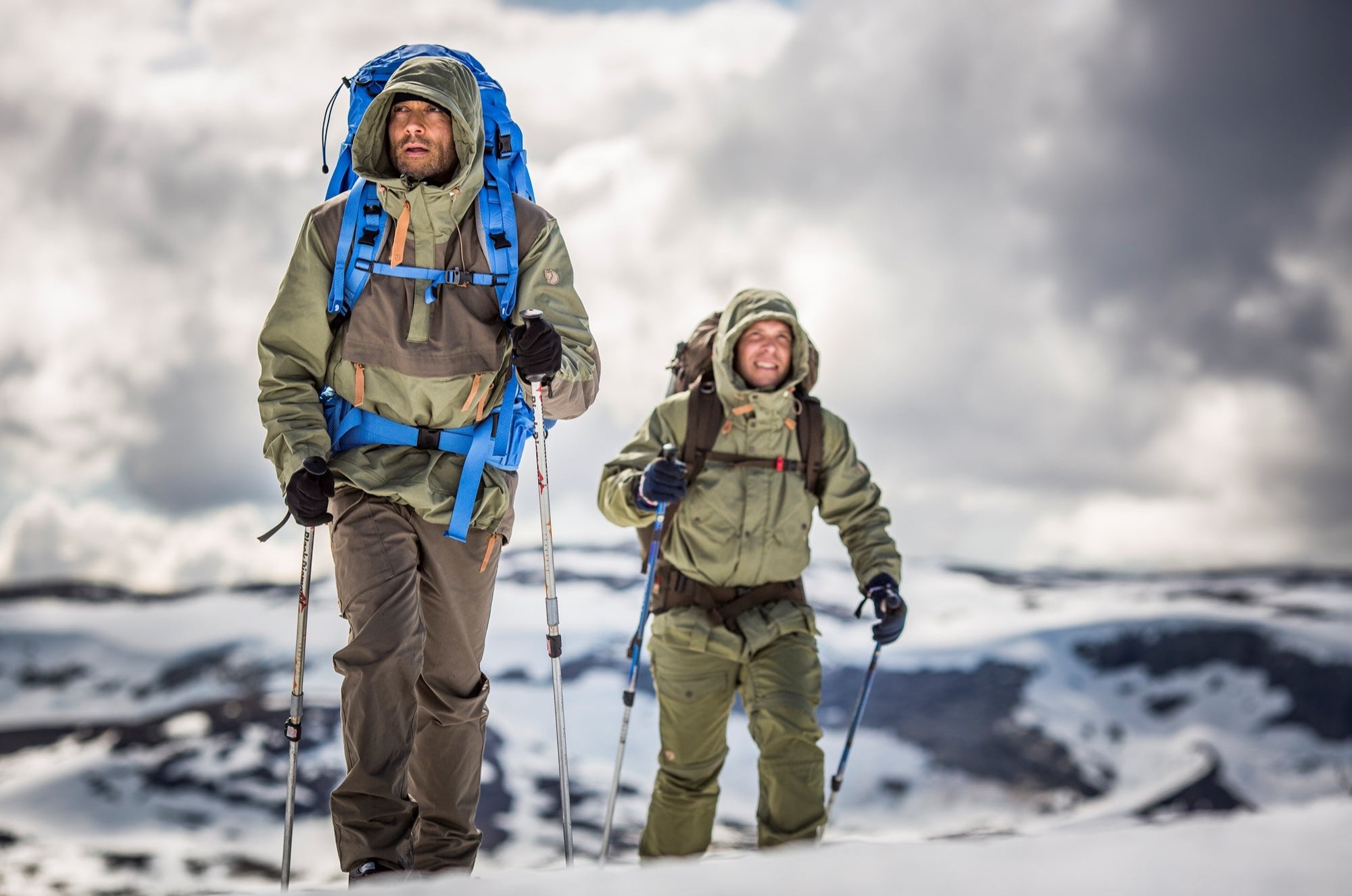 Two people in winter gear hiking on a snowy landscape with a cloudy sky.